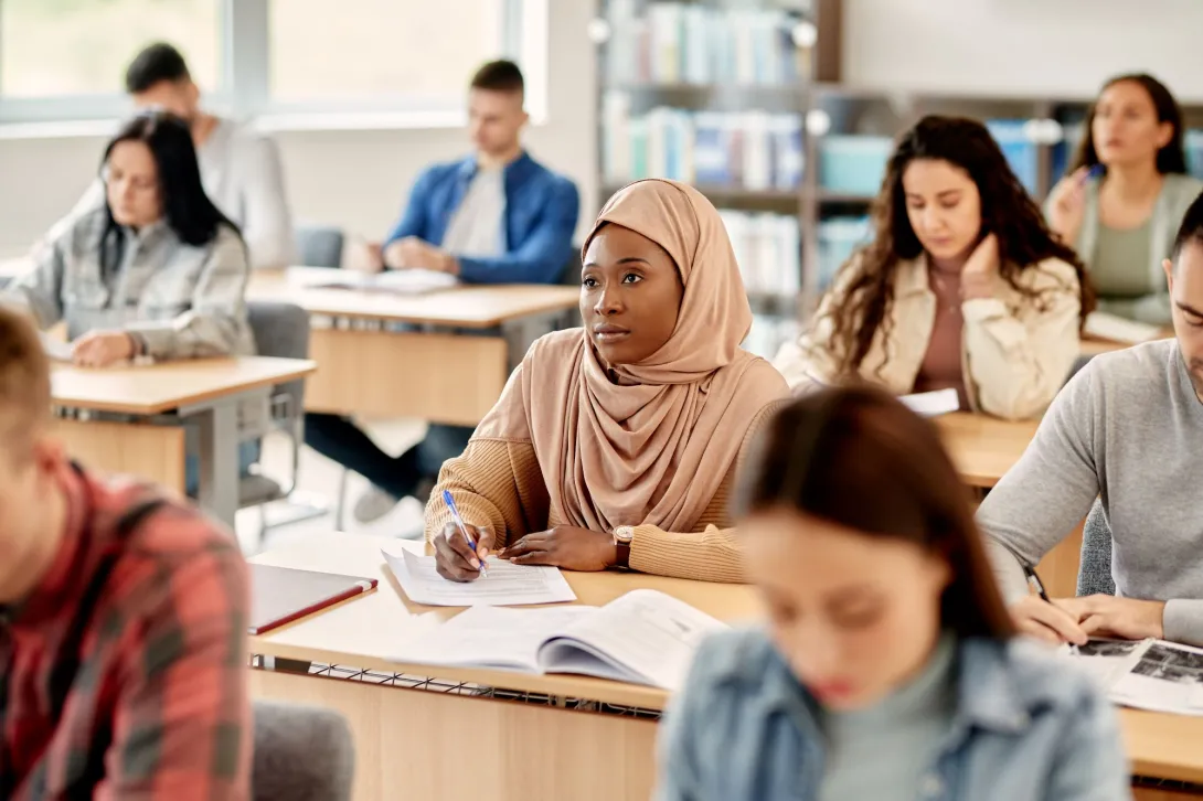 A female student wearing a hijab sits among other adult learners in a classroom.